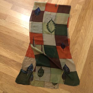 May include: A rectangular scarf with a patchwork design featuring squares of orange, brown, green, and beige. The scarf is adorned with leaf illustrations in shades of blue, green, and brown. The word "ECHO" is visible on the scarf.