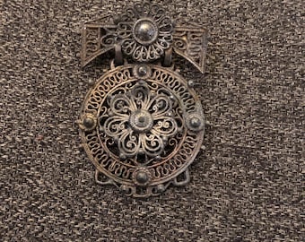 May include: An antique silver-toned brooch with intricate filigree detailing. The brooch features a circular design with a central floral motif and a decorative top piece. The metalwork showcases delicate patterns and textures, creating an ornate and vintage aesthetic.
