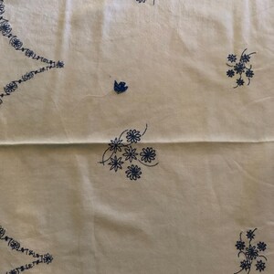 May include: Cream-colored fabric with embroidered blue floral patterns. The design includes clusters of flowers and a decorative border along the edges. The embroidery is detailed and evenly spaced across the fabric.
