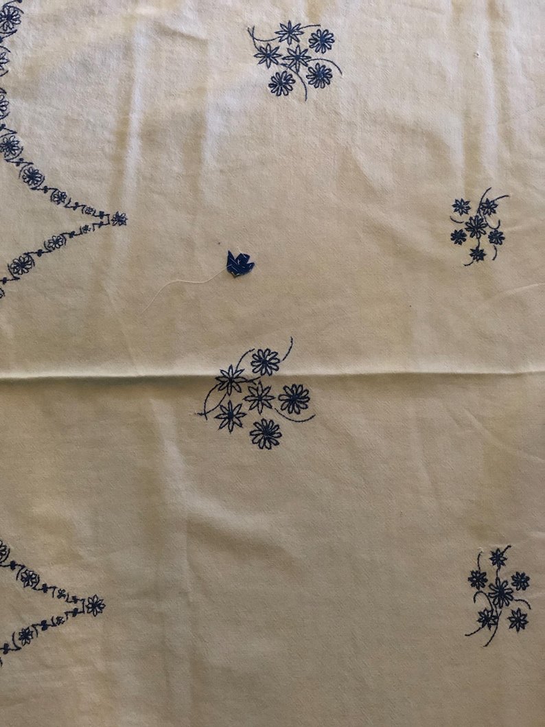 May include: Cream-colored fabric with embroidered blue floral patterns. The design includes clusters of flowers and a decorative border along the edges. The embroidery is detailed and evenly spaced across the fabric.