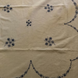 May include: Cream-colored fabric with embroidered blue floral patterns. The design includes scattered flower clusters and a scalloped border with repeating floral motifs. The fabric appears to be a tablecloth or similar textile.