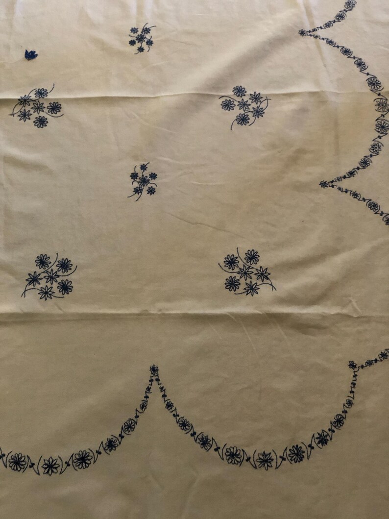 May include: Cream-colored fabric with embroidered blue floral patterns. The design includes scattered flower clusters and a scalloped border with repeating floral motifs. The fabric appears to be a tablecloth or similar textile.
