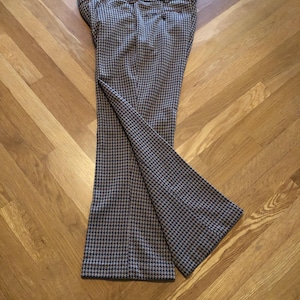 May include: Houndstooth pattern trousers in shades of brown, black, and white. The pants are made of a woven fabric and have a flared leg design. The trousers are laid out on a wooden floor.