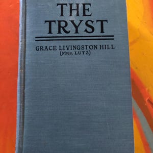 May include: A blue book with the title "THE TRYST" by Grace Livingston Hill (Mrs. Lutz).