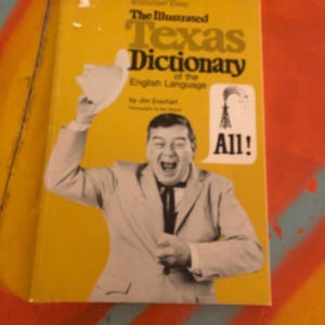 May include: A yellow book titled Volume Two The Illustrated Texas Dictionary of the English Language by Jim Everhart. The cover features a black and white photograph of a person in a suit and bolo tie waving a hat, with an oil derrick icon and the word All!