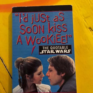 May include: A book cover with a blue background featuring a quote from Star Wars: "I'd just as SOON kiss A WOOKIEE!" The quote is in white text with a black outline. The cover also features a photo of Princess Leia and Han Solo looking at each other. The book title is "The Quotable Star Wars" in white text with a black outline. The author is listed as "Compiled by Stephen J. Sansweet".