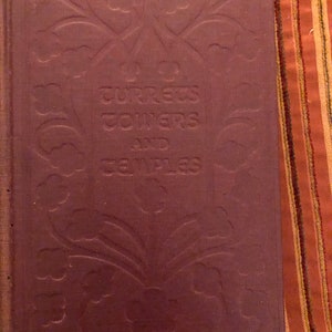 May include: A vintage book with a dark burgundy cover. The cover features embossed floral designs and the title "Turrets, Towers and Temples" in gold lettering. The book is closed and appears to be in good condition.