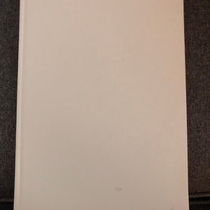 May include: A blank white book with a white cover and spine. The book is closed and lying flat on a gray surface.