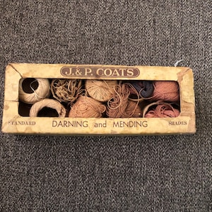 May include: A vintage box of J. & P. Coats darning and mending thread in various shades. The box is rectangular with a worn, aged appearance. Inside, there are multiple spools and bundles of thread in shades of brown, tan, and pink.