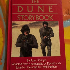 May include: A red book titled "The Dune Storybook" with a photo of two men in desert-like attire. The book is by Joan D. Vinge, adapted from a screenplay by David Lynch, and based on the novel by Frank Herbert.
