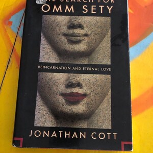 May include: A hardcover book titled "THE SEARCH FOR OMM SETY" by Jonathan Cott. The cover features a black background with two stone-like faces, one with red lipstick. The subtitle reads "REINCARNATION AND ETERNAL LOVE."