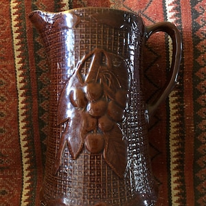 May include: A brown ceramic pitcher with a textured surface and a raised design of grapes. The pitcher has a handle and a wide mouth.