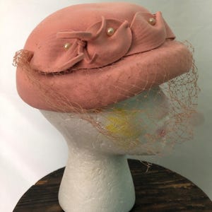 May include: A vintage peach-colored hat with decorative fabric accents and faux pearls. The hat features a delicate veil and is displayed on a white mannequin head. The hat is a classic accessory.