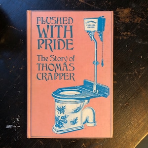 May include: A pink book titled "Flushed With Pride: The Story of Thomas Crapper" with an illustration of a blue and white toilet on the cover. The toilet has a floral design and a blue tank with the text "T Crapper Chelsea London".