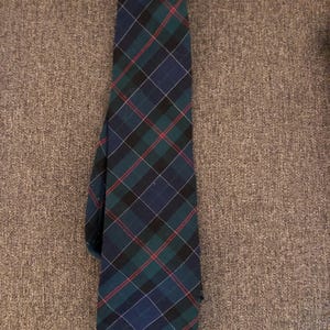 May include: A navy blue, green, black, and red plaid tie. The tie features a classic tartan pattern with intersecting lines and a pointed end. The tie is made of a woven fabric and is suitable for formal or business attire.