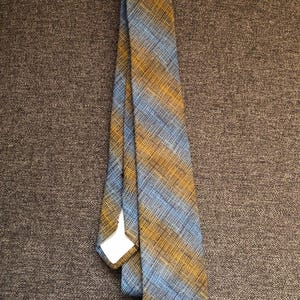 May include: A necktie with a diagonal pattern in shades of blue and gold. The tie is made of a woven fabric and has a classic shape. The tie is displayed on a brown textured surface.