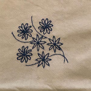 May include: Close-up of a cream-colored fabric with a cluster of embroidered blue flowers. The flowers have detailed petals and are connected by thin, curved stems. The embroidery adds a delicate, handmade touch to the textile.