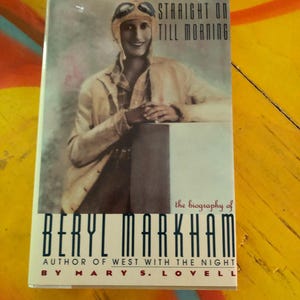 May include: A book cover featuring a vintage photograph of a person wearing a flight helmet and goggles. The title reads "STRAIGHT ON TILL MORNING" above the biography of Beryl Markham. The author's name is Mary S. Lovell.