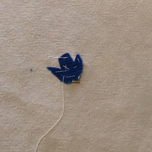 May include: A close-up of a navy blue felt flower with white thread stitching. The flower is attached to a beige fabric background. The thread extends from the flower, suggesting it is being sewn.