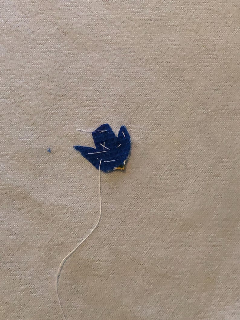 May include: A close-up of a navy blue felt flower with white thread stitching. The flower is attached to a beige fabric background. The thread extends from the flower, suggesting it is being sewn.