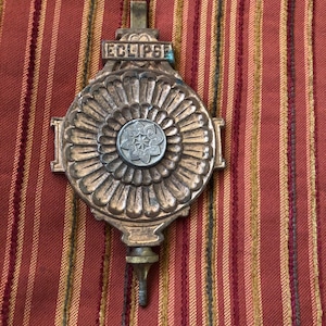 May include: A metallic clock pendulum featuring a circular, fluted design with a central silver-toned floral medallion. The word ECLIPSE is embossed on a rectangular plate at the top. A threaded adjustment screw extends from the bottom.