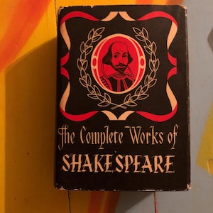 May include: A black book with a red and white border. The cover features a portrait of William Shakespeare with a laurel wreath around it. The title "The Complete Works of SHAKESPEARE" is printed in white on the cover.