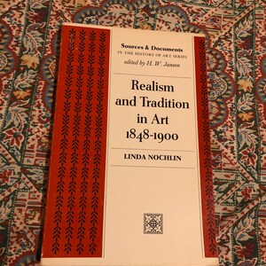 May include: A book titled "Realism and Tradition in Art 1848-1900" by Linda Nochlin. The cover is white with red accents and text. The book is part of the "Sources & Documents in the History of Art Series".