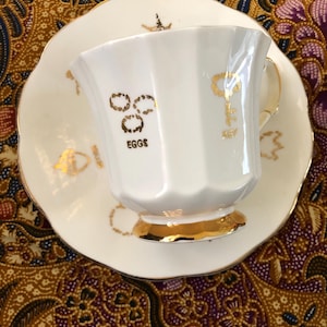 May include: White teacup and saucer set with gold trim and accents. The teacup has a unique shape and features the word "EGGS" in gold lettering. The saucer has a scalloped edge and gold detailing.
