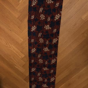 May include: A long, rectangular scarf with a burgundy background. The scarf features a repeating pattern of dark blue leaves and white grape clusters. The edges of the scarf are a darker burgundy.