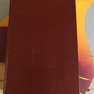 May include: A closed, rectangular book with a dark reddish-brown cover. The cover appears to be made of a smooth material. The book is positioned vertically, taking up most of the frame. The background is a light gray with orange and yellow accents.