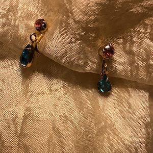 May include: A pair of gold-toned earrings. Each earring features a small, round, pink gemstone at the top and a teardrop-shaped, blue gemstone dangling below. The earrings are set against a textured, golden-colored fabric.