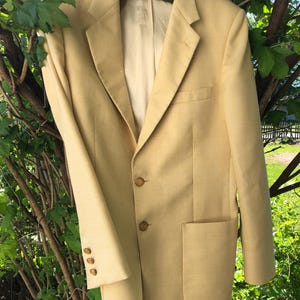 May include: A light yellow blazer with a two-button closure and three buttons on each sleeve. The blazer has a notched lapel, a chest pocket, and two lower pockets. The jacket is hanging on a green hanger.