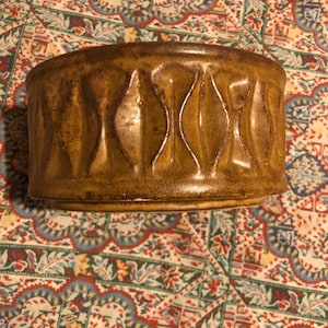 May include: A cylindrical, ceramic container with a textured, brown glaze. The container features a repeating pattern of vertical, elongated diamond shapes. The piece is set against a patterned fabric background.