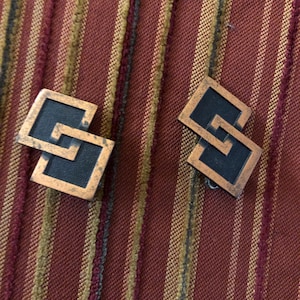 May include: Two vintage copper-toned square cufflinks with a black center and a geometric design. The cufflinks are displayed on a striped maroon, gold, and olive green fabric background.