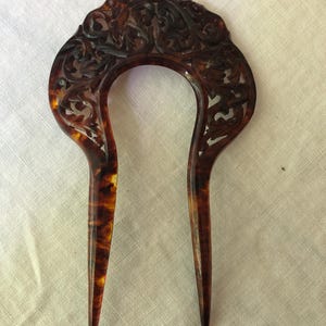 May include: A vintage hair comb with a U-shaped design. The comb is made of a tortoiseshell material with intricate carved floral patterns. The comb's color is a mix of brown and amber.