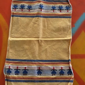 May include: A rectangular yellow woven textile featuring a fringed edge at the top and bottom. The fabric displays horizontal stripes in blue, red, and white, accented by a repeating pattern of small blue and red figures across the top and bottom.