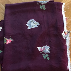 May include: Burgundy fabric with a floral rose pattern. The roses are white, pink, and red with green leaves. The fabric is folded, showing the raw edge of the material.