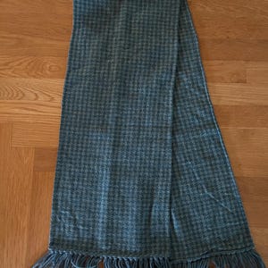 May include: A rectangular scarf featuring a blue and grey houndstooth pattern throughout the fabric. The scarf has a textured appearance and is finished with matching blue and grey fringe detailing along both of the shorter ends of the garment.
