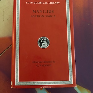 May include: A red hardcover book titled Loeb Classical Library Manilius Astronomica, edited and translated by G. P. Goold. The cover features a white Greek key border, white text, and a circular black and white emblem of a seated figure.