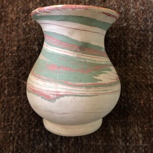 May include: A small, white ceramic vase with a marbled design in shades of green and pink. The vase has a rounded body and a flared opening.