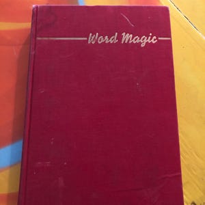 May include: A red book with the title "Word Magic" in gold lettering. The book has a simple, classic design, with a solid red cover and a gold horizontal line above the title. The book appears to be in good condition.
