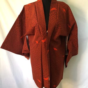 May include: A rust-colored kimono with a geometric pattern and orange leaf-shaped accents. The kimono is displayed on a black mannequin and has wide sleeves and a draped front. The fabric appears to be textured.