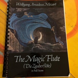 May include: A spiral-bound music score titled "The Magic Flute" by Wolfgang Amadeus Mozart. The cover features a dark illustration with figures in flowing blue robes, a crescent moon, and the text "The Magic Flute (Die Zauberflöte) in Full Score."