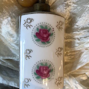 May include: A vintage perfume bottle with a white background, featuring two pink rose designs within gray dotted circles. The bottle has gold accents and a tan bulb atomizer.