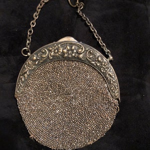 May include: A vintage silver beaded purse with a decorative floral frame and a chain handle. The round purse is covered in small, shimmering beads. The frame has an ornate floral design. The chain handle is attached to the frame.