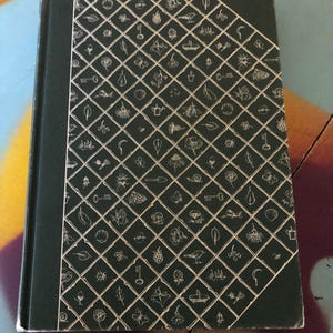 May include: A hardcover book with a dark green cover and spine. The cover features a white grid pattern filled with small, hand-drawn illustrations of keys, flowers, and other natural elements. The book's edges show slight wear.