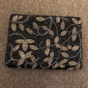 May include: Black beaded clutch with silver and gold leaf design. The rectangular evening bag features a detailed floral pattern. The beaded embellishments create a shimmering effect, perfect for special occasions.