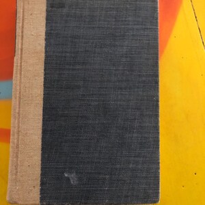 May include: A black hardcover book with a tan spine. The book is lying on a yellow and orange surface.