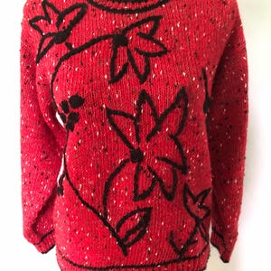 May include: A long-sleeved knit sweater in a vibrant red speckled yarn with black flecks. The sweater features a prominent black embroidered floral pattern across the front, with a solid black ribbed crew neckline, cuffs, and hemline.