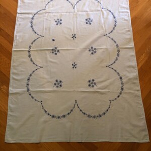 May include: A white tablecloth with a large, scalloped border and floral embroidery in blue. The embroidery features clusters of small flowers and a delicate, repeating pattern along the edge. The tablecloth is rectangular and appears to be made of a soft fabric.